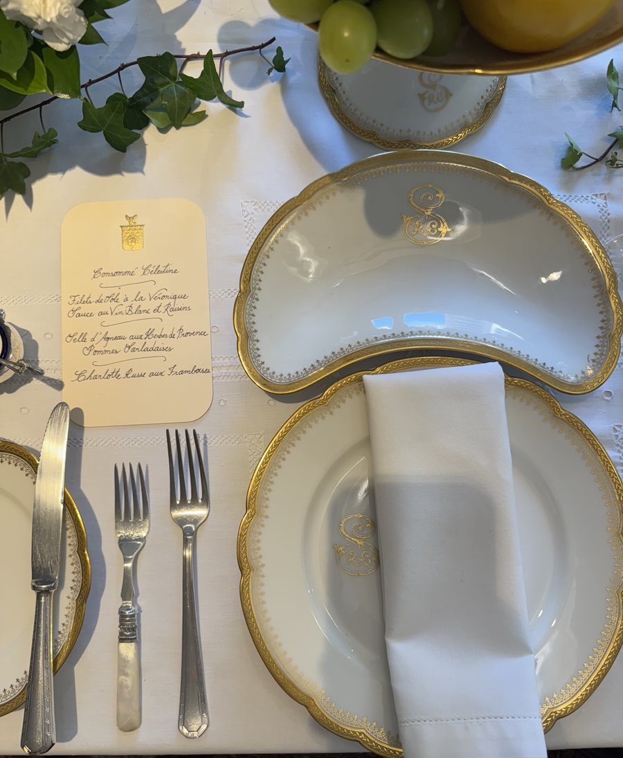 Um lugar à mesa visto de cima: pratos brancos com borda e monograma dourados, guardanapo dobrado, talheres de prata e um menu escrito à mão, com brasão dourado.