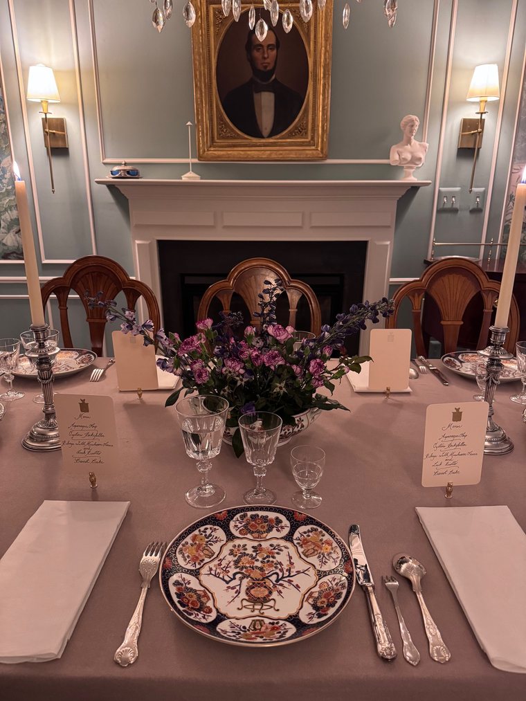 Mesa posta numa sala de paredes claras com lareira e retrato: prato de porcelana decorada, garfos à esquerda, faca e colheres à direita, castiçais, flores e menus escritos à mão.