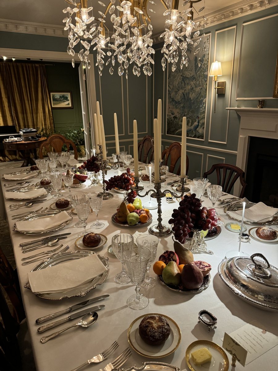 Mesa de jantar longa posta à luz de velas, com candelabros de prata, cristais, frutas e um lustre de cristal, numa sala de paredes verdes.