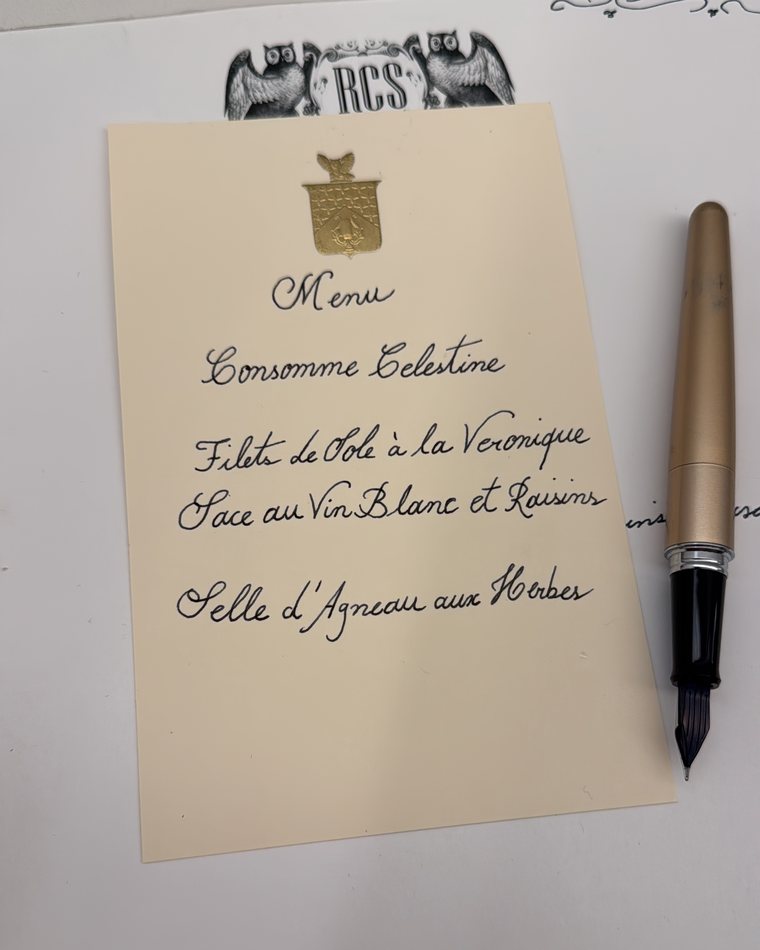 Um menu escrito à mão em papel marfim, com brasão dourado, sobre o papel timbrado de Rafa com as corujas e o monograma RCS, ao lado de uma caneta-tinteiro.